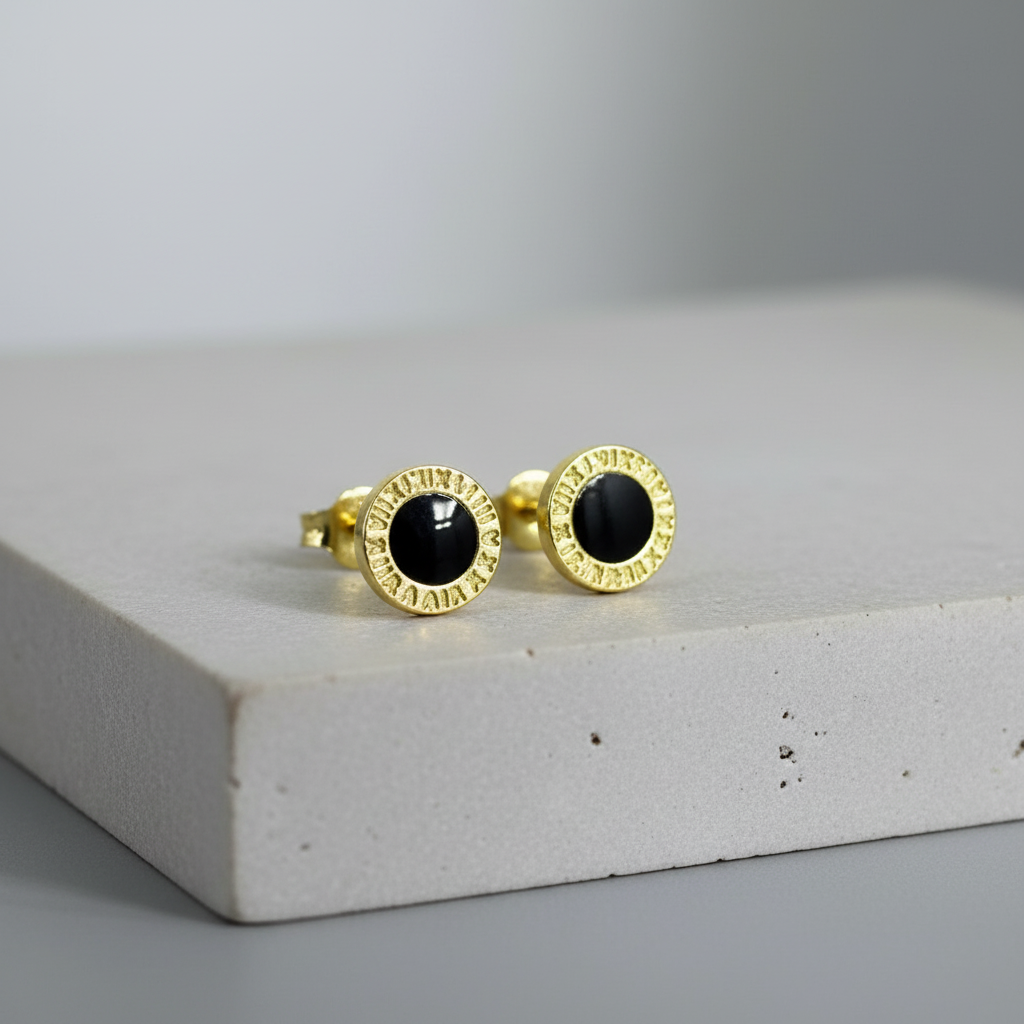 Gold-rimmed black enamel stud earrings on grey stone podium